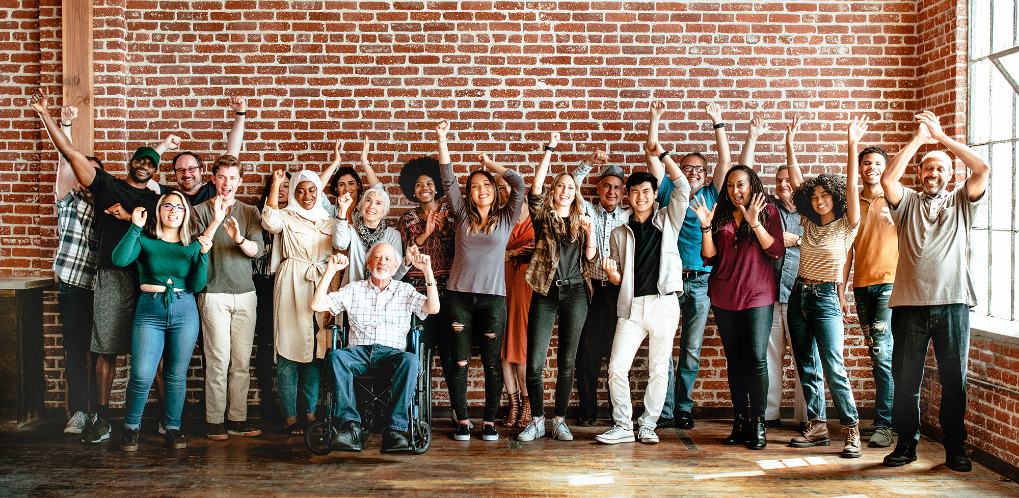A diverse group of people in front of a brick wall with their arms raised in celebration.
