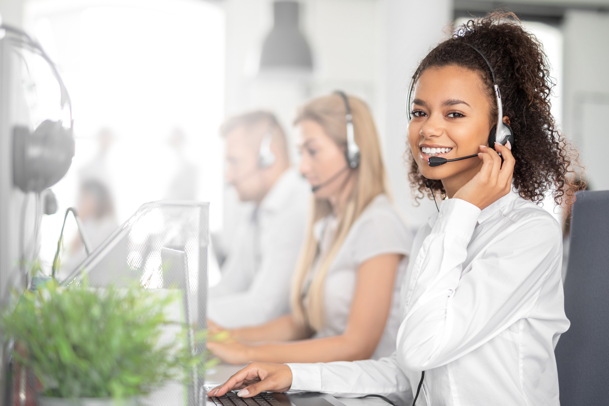 A customer service representative in a headset sits at in front of aa keyboard. She is looking into the camera and smiling.