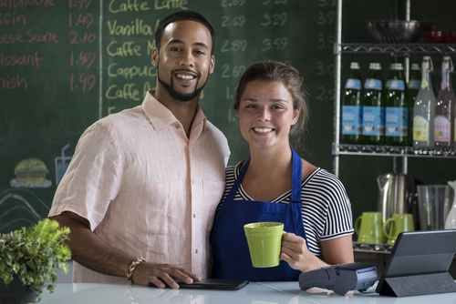 Un hombre y una mujer de pie detrás del mostrador de una cafetería. Están sonriendo para la cámara.